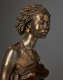 Bronze bust of a woman with poised expression, raised chin, hair in twists, gold earrings, beaded necklaces accentuating her exposed collarbones and bare shoulders above a strapless tightly-wrapped dark top.