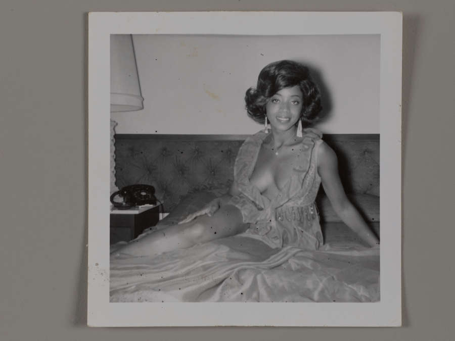  Black and white vintage snapshot of a smiling young dark-skinned woman. She sits on a sofa in a negligee and dangling earrings, cleavage exposed. She looks directly at the camera. 