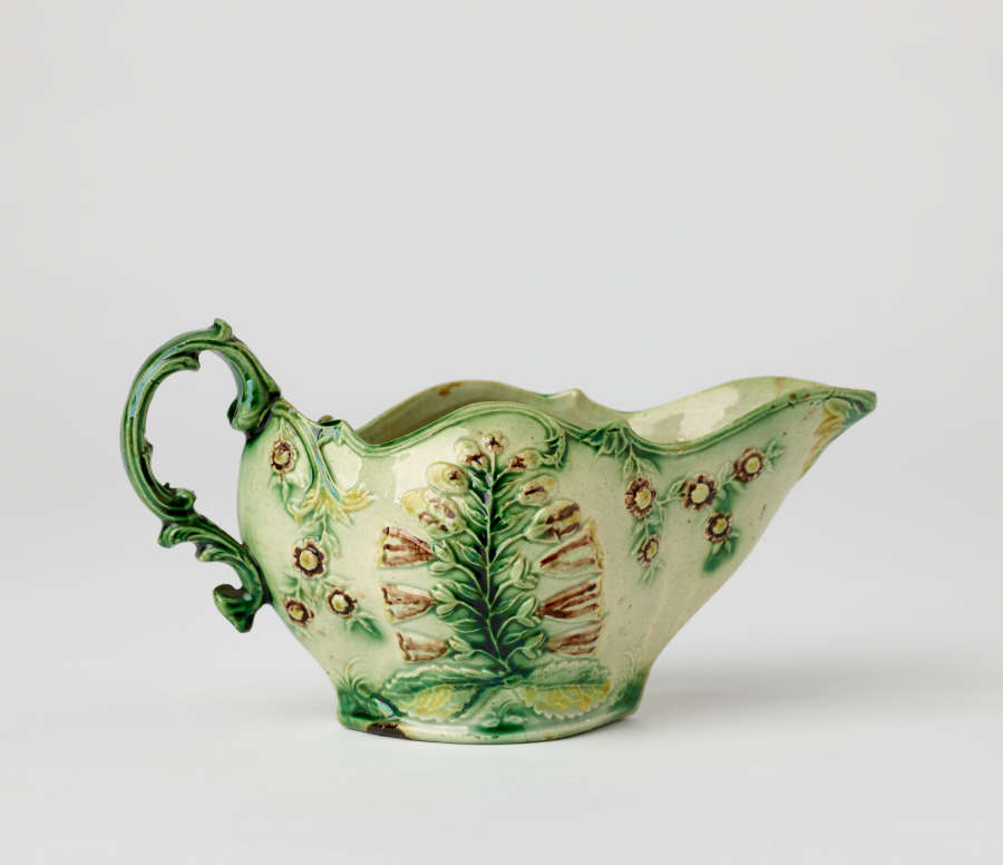  A short boat shaped pitcher green, cream, and brown in color with a sculptural handle and floral decorations.
