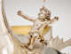 Detail of silver cherub gazing upwards with outstretched arms wearing a short, draped cloth, kneeling with one knee up at the base of a curved silver surface. 