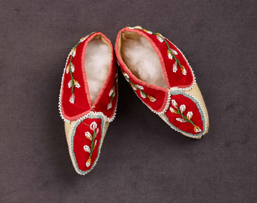 Top view of a pair of red moccasins with white and green floral embroidery and tan soles. The insides of the moccasins are stuffed with a white cotton-ball like material.