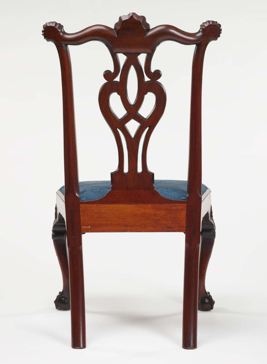 Rear view of a wooden chair with an intricately carved back and an upholstered blue seat supported by slender legs.