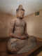 An aged wooden Buddha statue seated in meditation on a raised platform in a warmly lit room. The statue sits cross-legged in a meditative pose with a serene expression.