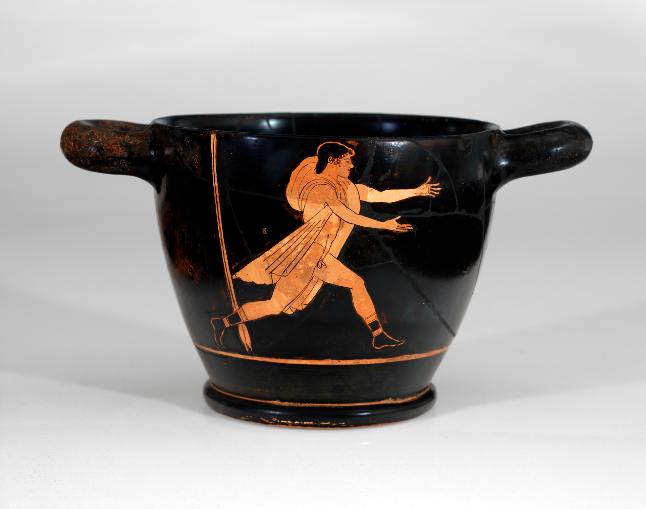 Rounded black vessel with thick handles and a cracked surface featuring orange illustrations of a partially nude figure wearing draped fabric besides a tall horizontal arrow  running with arms outstretched.