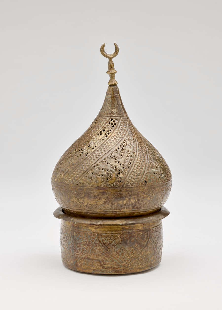 Bronze-colored sculptural object with incised design. Top half is teardrop shaped with a crescent moon shape on top. Cylindrical vessel on bottom.