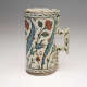 A rounded cream colored vessel with a square handle, decorated with geometric and flower designs in blue, green, and red.