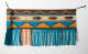 Turquoise, yellow, and beige woven fabric with fringed ends, geometric patterning, and thin tassels at the top corners. The left side of the fabric is rolled, shortening its length. 