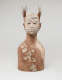 Brown clay bust of a figure with a grass and stick headdress. Diagonal lines of patterned patches of offwhite run across their chest and cover their face.