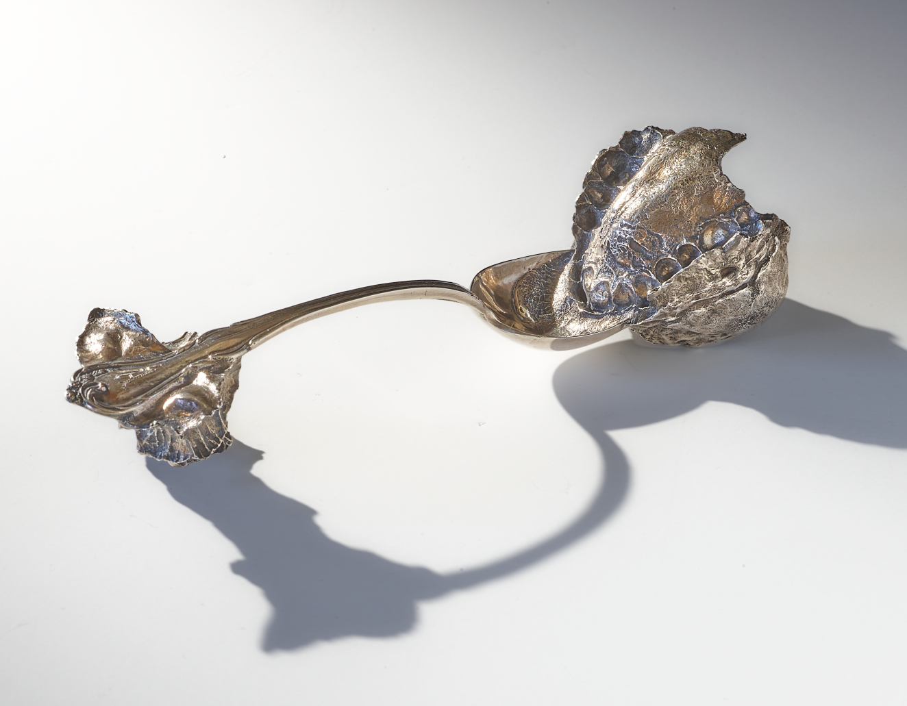 Silver spoon altered roughly at each end. Attached to the bowl is a clump that has been deeply bitten by a human mouth. The handle bears a smaller jagged form.