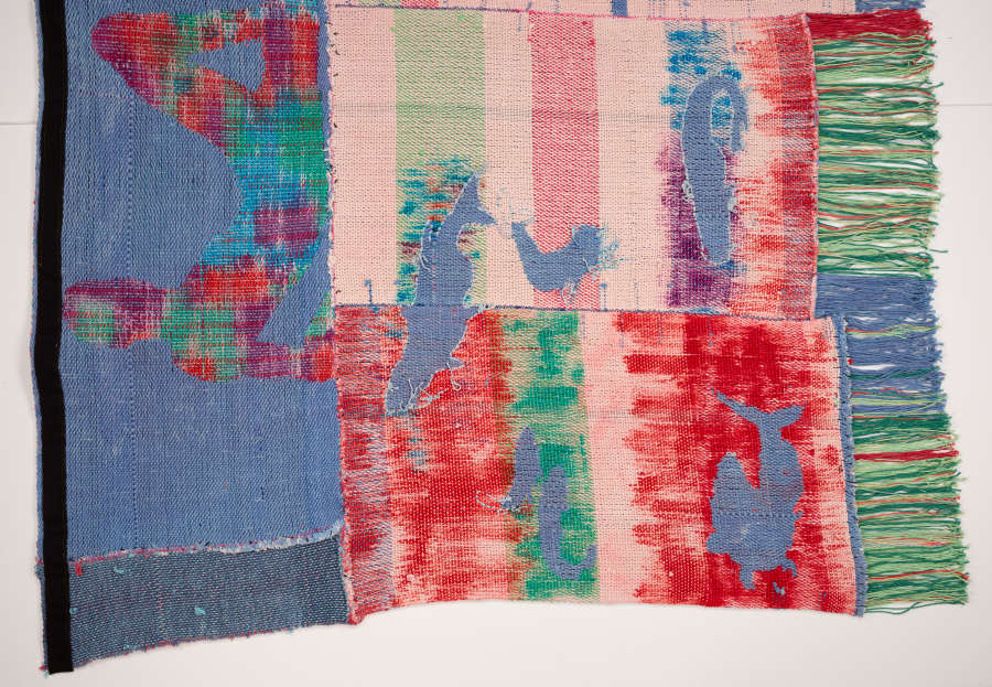 View of half of the backside of the tapestry. One edge is frilled. This end features an inverse of the font with silhouettes previously blue, now patterned and vice versa.