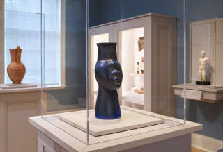 In a case in a gallery’s center is a sleek black vessel shaped like a stylized head with an elongated neck. Cases along the perimeter contain other objects.