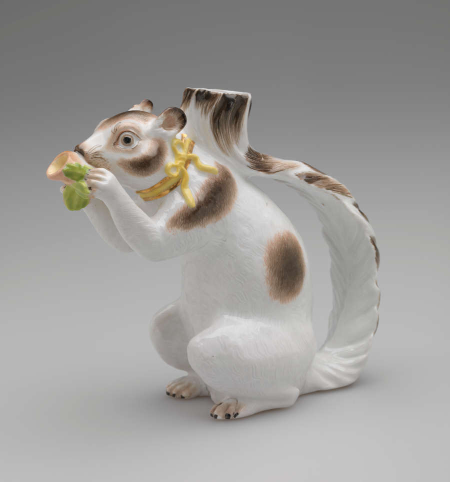 Vessel in the shape of a white squirrel with brown patches and a yellow ribbon arounds its neck. It holds a brown nut with green leaves up to its face.