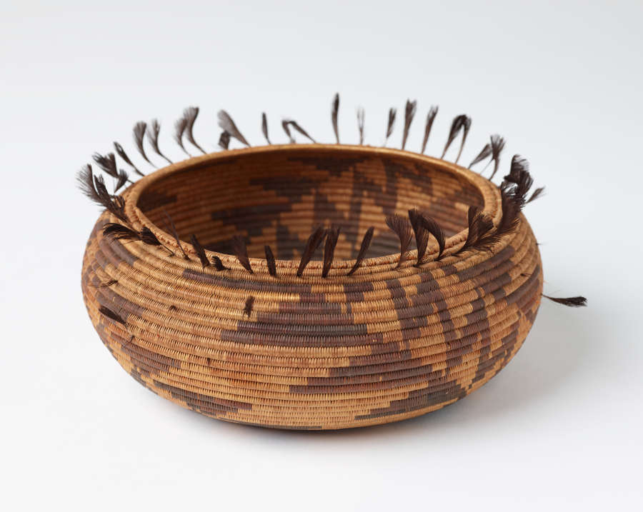Woven tan and brown onion shaped bowl with geometric dark brown patterning and feather-like tassels emerging from the top edge of the bowl.