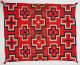 Red woven cloth with red-centered, geometric cross-shaped patterns throughout and red tassels in the corners. The navy blue and cream bordered crosses alternate between cream and navy blue.