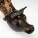Close-up of a black ceramic vessel shaped like a bull’s head with alert eyes, short horns, and a red, muscular figure painted on its neck, framed by decorative patterns. 