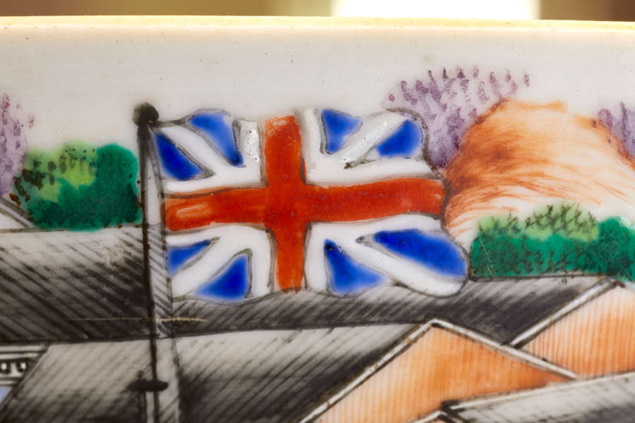 A close-up of a painted ceramic depicting the British flag on a pole, positioned above a building with green foliage and hills in the background.