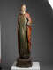A tall, weathered wooden statue of a robed figure with a serene expression, draped in green and red garments, standing upon an octagonal base.