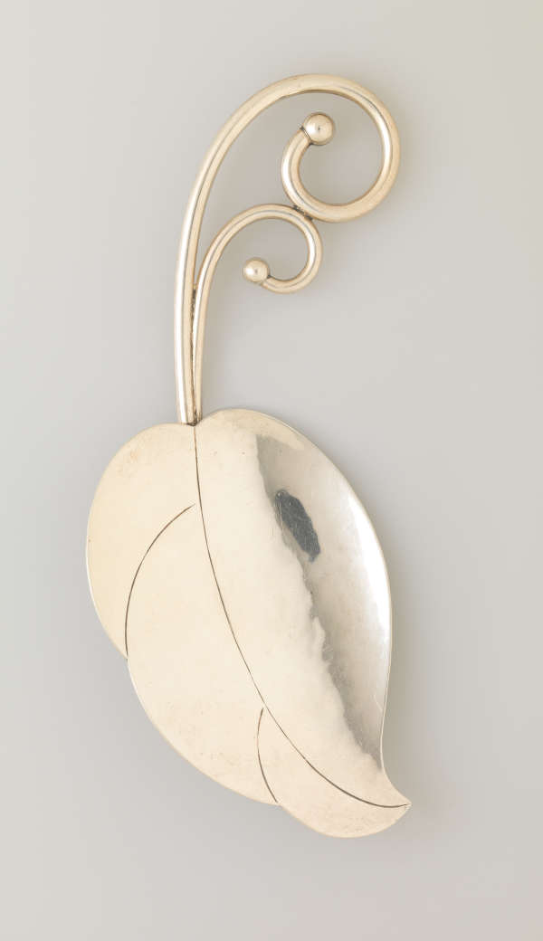 A leaf shaped ornament made of silver with two spiraling stems emerging from the top. On the right edge is a polished section which reflects the gray backdrop.