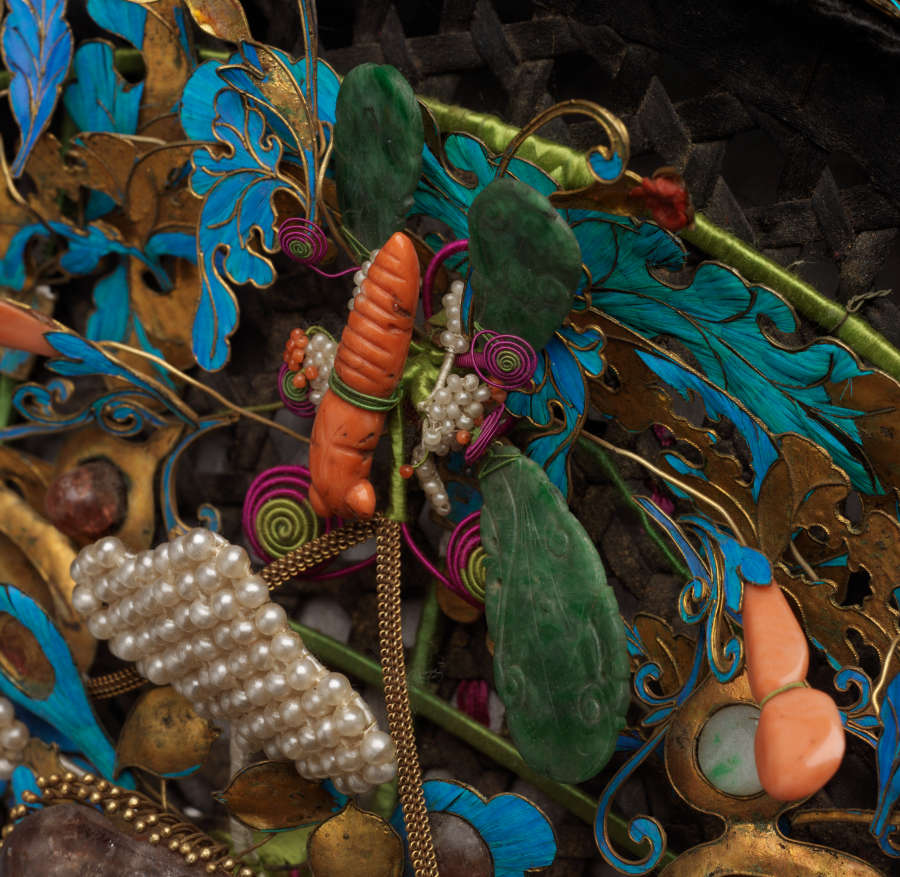 Angled close-up of the headdress showing the stacked layers of elements on the surface of the headdress. Visible are electric-blue leaf ornaments and an orange stone insect with jade wings.