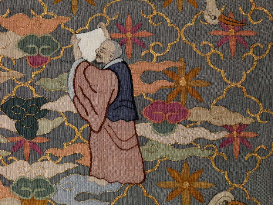 Robe’s back detail, showing a standing robed monk looking upwards, holding a scroll. The background is dark with a thin golden diagonal grid, within each square is a floral motif.