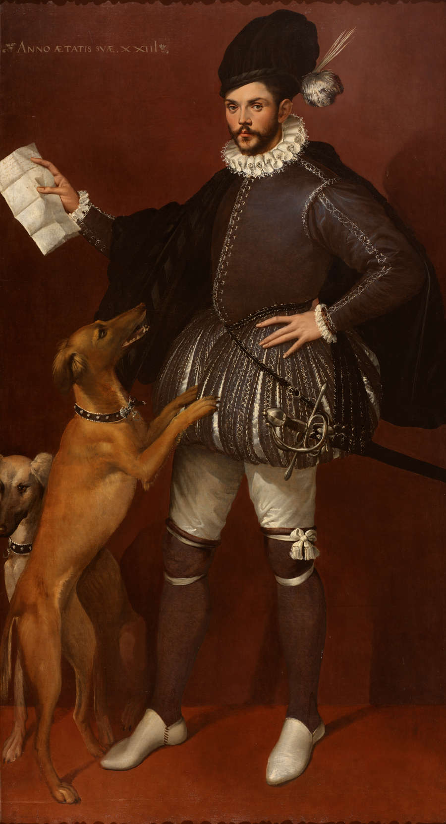 A lavishly dressed, bearded, European gentleman holding up a letter in his left hand. He has a sword at his right side and is ignoring two dogs.