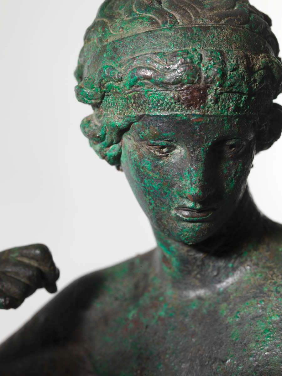 Detail view of a bronze statue of a crowned nude woman at an angle. Visible are the details of her face, hair and clenched fist curled towards her shoulder.