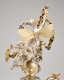 Back view of a silver-robed figure with flowing hair and thin, gold, insect-like, leaf-textured wings holds a draping garland aloft with two cherubs atop a gold orb. 
