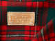 Close-up of a tan rectangular label with orange embroidered text that reads “warranted to be a ‘Pendleton.’ Pendleton Woolen Mills. Portland, Oregon 100% Virgin Wool, ” on a red and green plaid long-sleeved shirt. 