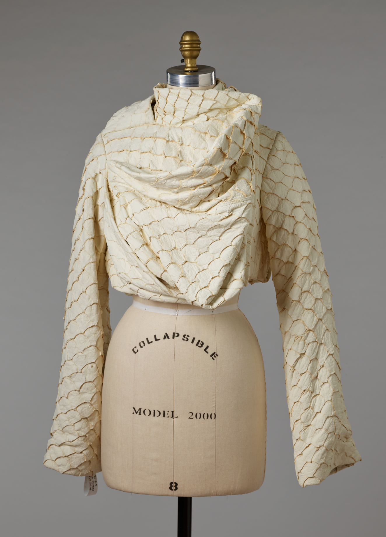 A cream colored, draped garment with wide sleeves and gold-accented scallop designs, fitted on a mannequin torso.