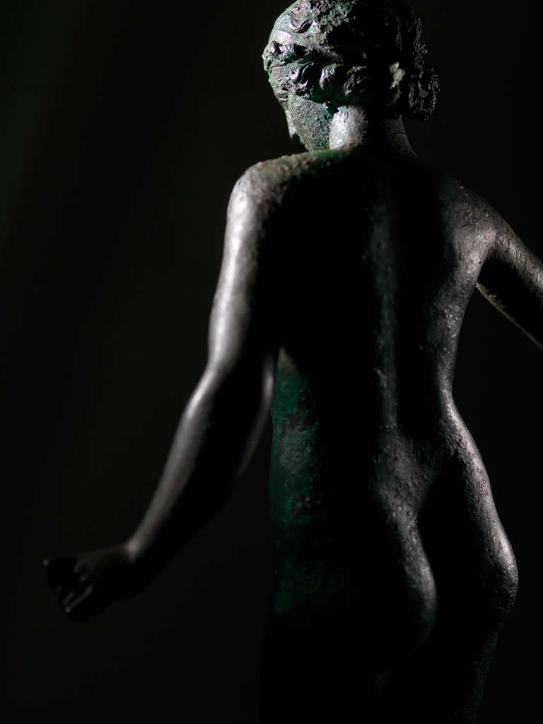 Back view of a tarnished bronze statue of a standing nude woman, her arms away from her body, as she emerges from the darkness. The lighting amplifies the texture of the statue’s surface.