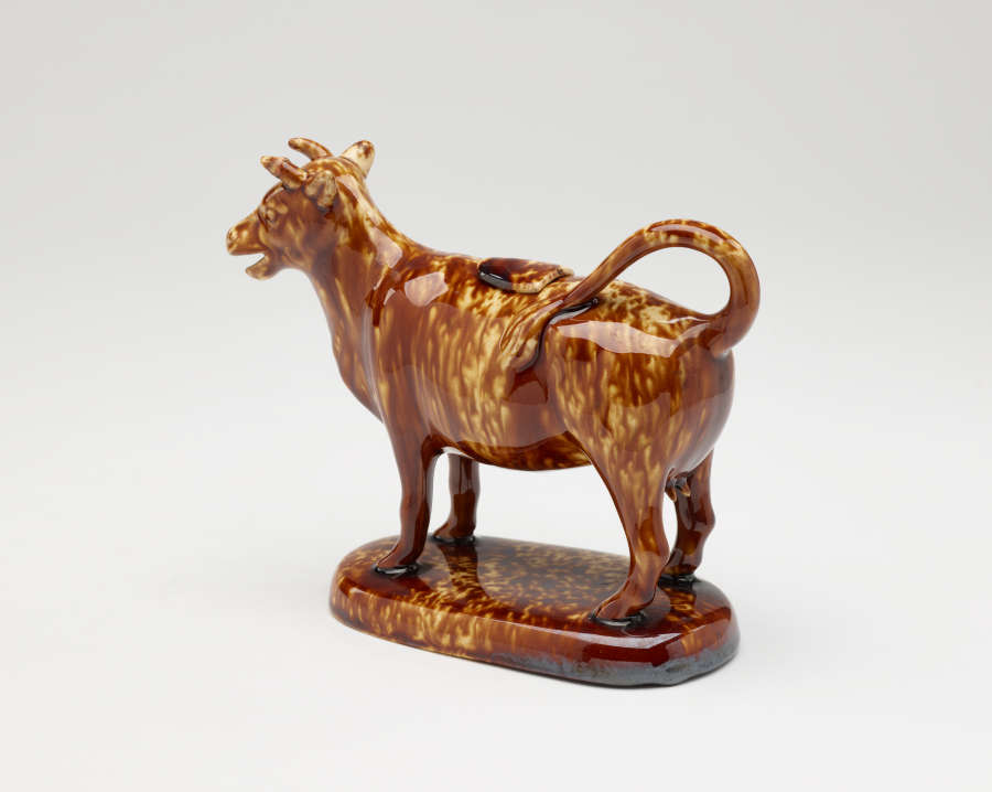  A brown sculptural creamer in the shape of a cow with horns. There is a small flat lid in the center of its back.