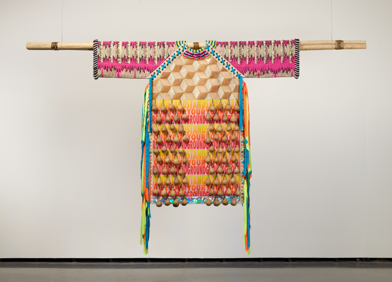 A garment displayed on a wooden pole, with bright woven geometric patterns, dangling gourds, and repeated pink text reading "STAND YOUR GROUND" woven into the textile.