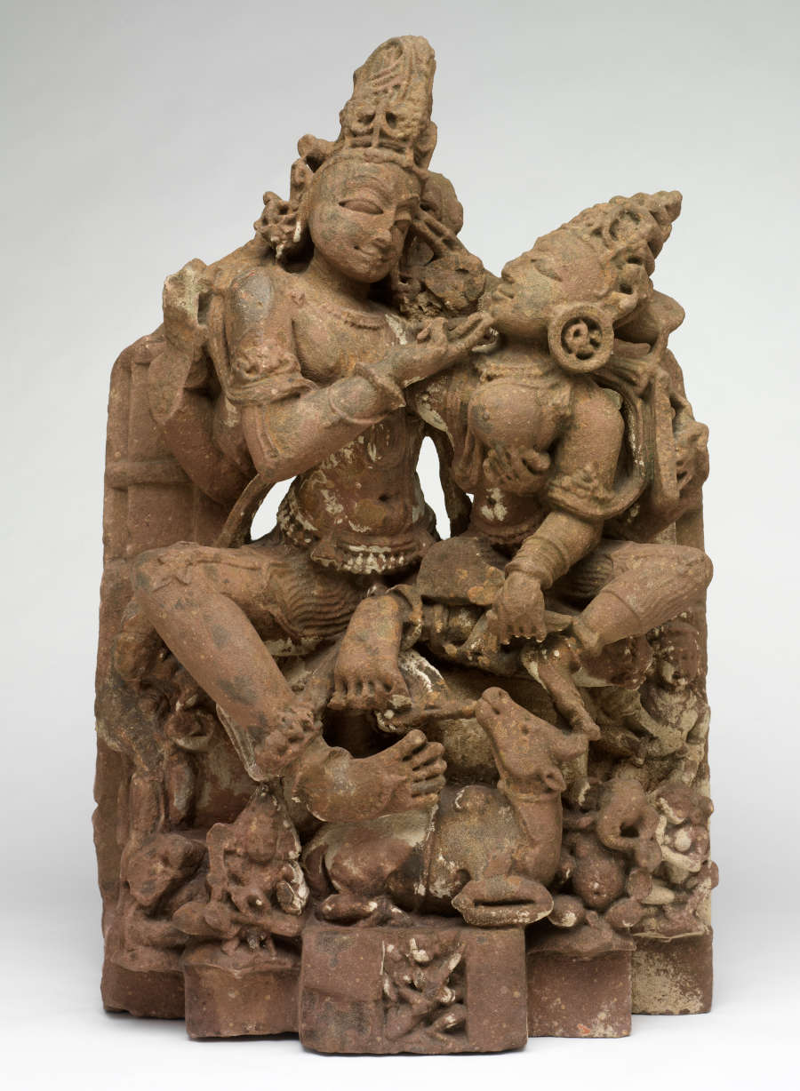 Brown sculpture of two figures caressing each other. Both figures wear headdresses and minimal clothing. Various animals watch from below, and they are all situated on a platform.