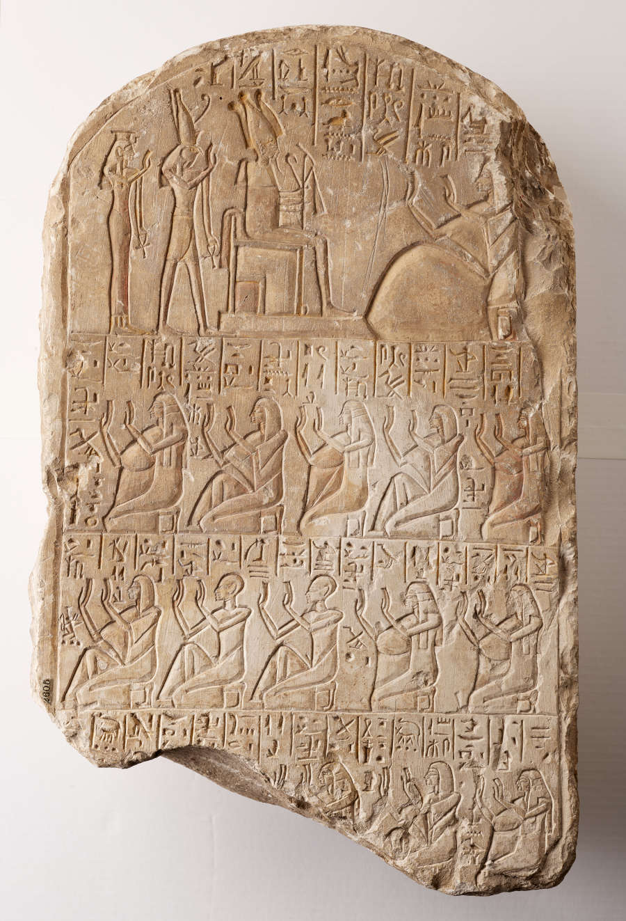 Thick weathered brown-gray long arched stone fragment against a wooden-textured background. The fragment features rows of raised lines forming figures kneeling and featuring towards one another, along with scripture.