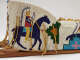 Detail of beaded object with leather ties hanging down and wood supports at bottom. White background beaded with blue horse and colorful rider in headdress, with geometric decoration.  