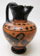 Angled view of an onion-shaped black and orange jug with a pinched mouth and handle, along with illustrations of a sphinx, roosters, and floral motifs painted on the surface.