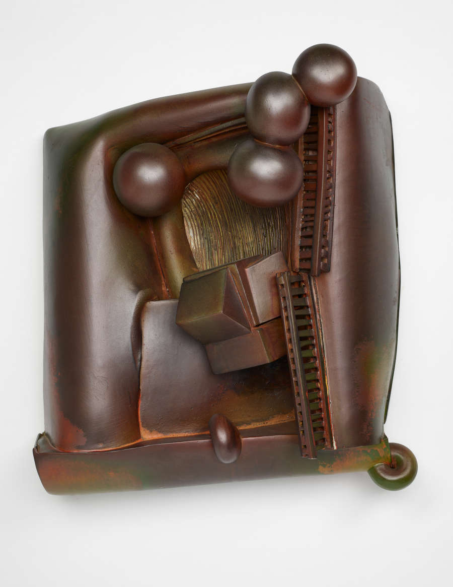 A rectangular, metallic bronze-colored abstract sculpture. The surface is uneven, with small, round globes and multifaceted, angular shapes nestled into an indentation in the center resembling a couch.