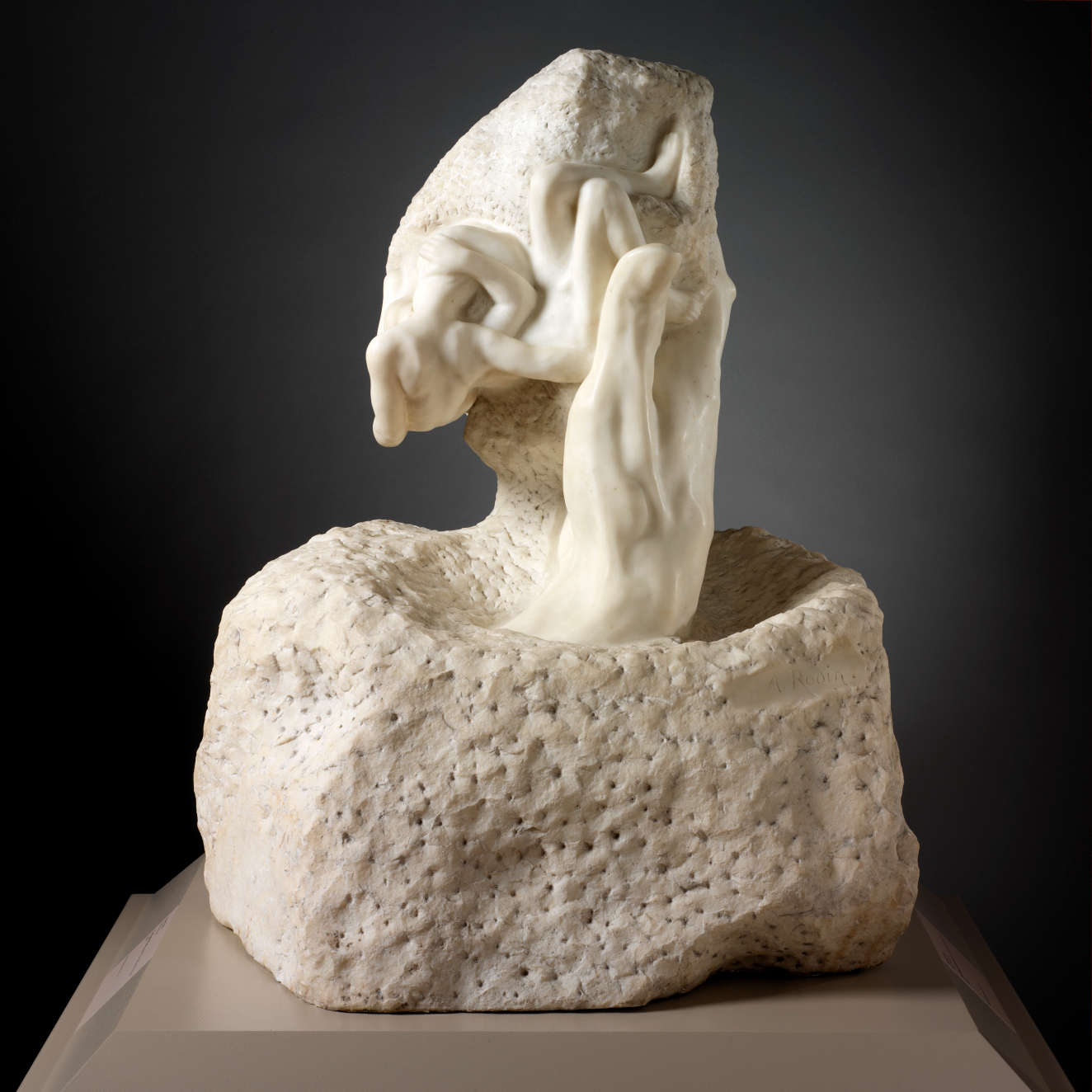 Marble sculpture depicting the thumb of a carved hand holding rock from which two emerging figures flow into each other,
rising from a base of unworked stone. 