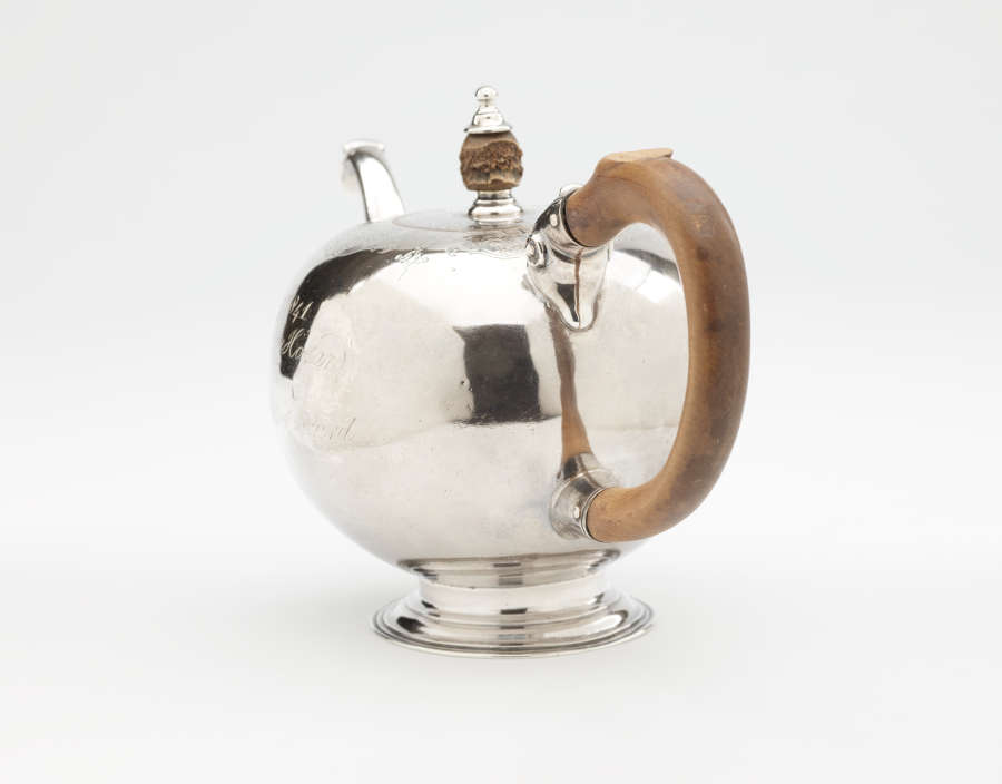 A bulbous silver teapot with a wooden finial and handle connected with decorative silver pieces. There are delicate engraved marks on the body and lid.