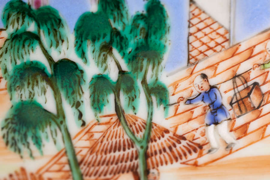 A close up of a painted ceramic depicting a figure in blue using a shoulder pole to carry a box, descending an angled orange brick-way with lush green trees in the foreground.