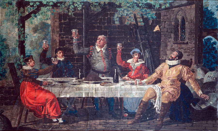 Jovial scene with figures in classical attire sat around a table toasting with goblets. The gathering is outdoors in a vineyard with a brick wall framing the table.