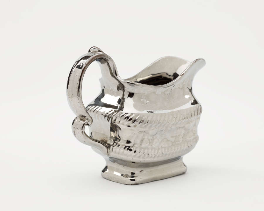  A silver creamer with a decorative handle and a rounded square body with sculptural decorations.
