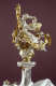 Robed silver figurine with flowing hair and thin gold wings holding a draped gold garland aloft, flanked by cherubs blowing horns atop an orb-like pedestal against a dark background. 
