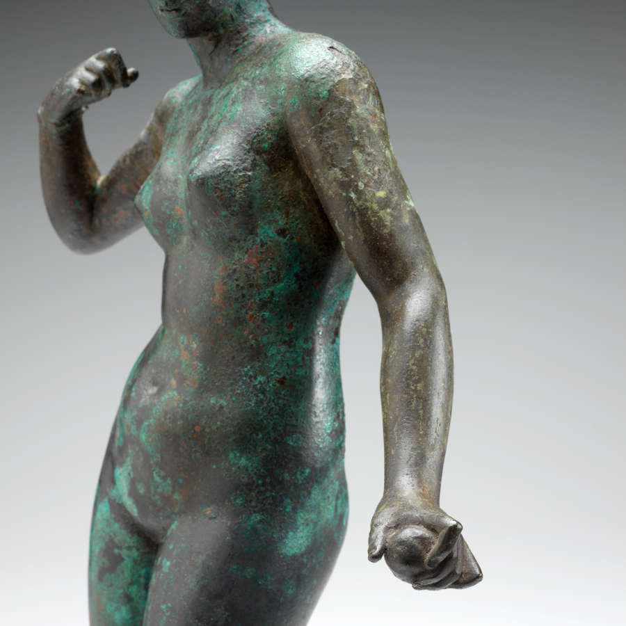 Gray metal sculpture cropped at the torso, with a green patina of a standing crowned nude woman, one arm down, grasping an object, and the other curled towards her shoulder.
