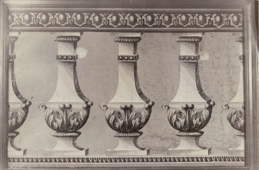 Black and white photo of repeated vases with raised leaf motifs lining its body and dotted embellishments decorating its neck. A floral, geometric border surrounds the vases on top. 