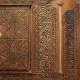 A close-up of a carved wooden panel with ornate calligraphy and symmetrical star patterns, bordered by intricate scrollwork and geometric motifs. 