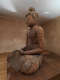 An angled view of an aged wooden Buddha statue seated in meditation with visible cracks and repairs sat in a warmly lit room.