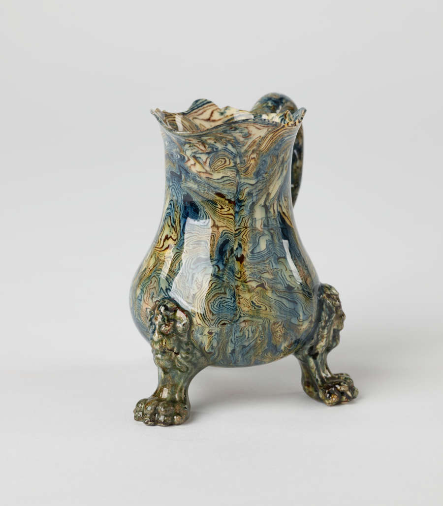 A small vessel with a cream, blue, and green mottled design. Three sculptural feet are shaped like lion’s paws with lion heads attached to body of vessel.