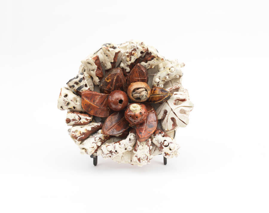 Circular ceramic sculpture featuring a cluster of glossy brown, seed-like pods at the center, surrounded by textured cream leaves speckled with brown radiating outwards from the cluster of seeds.
