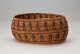 Side-view of an ochre-brown oval woven basket with irregular edges and convex sides featuring two patterned stripes of dark-brown figures holding hands separated by horizontally running stripes.